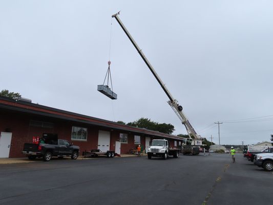 A white mobile crane on a street extending its boom to lift a rectangular mechanical unit toward a commercial building.