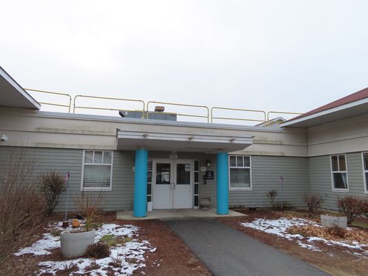 Exterior entrance of a grey one-story commercial building with blue supporting pillars and a concrete walkway.
