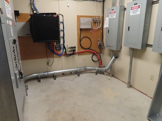 A mechanical room corner featuring a wall-mounted control box, vertical grey electrical panels, and metal conduits running along the floor.