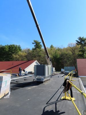 A rooftop view showing a crane lowering a large HVAC unit onto a flat roof where other equipment is already installed.