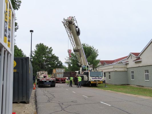 A wide shot of a crane positioned on a street next to a building, extending its boom to reach the roof.