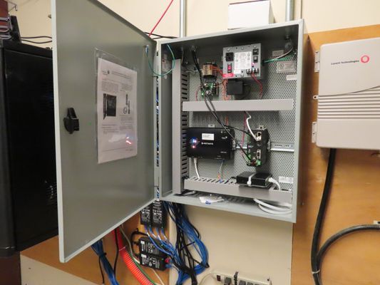 An open grey electrical control cabinet mounted on a wall, showing internal circuit boards, complex wiring, and communication modules.