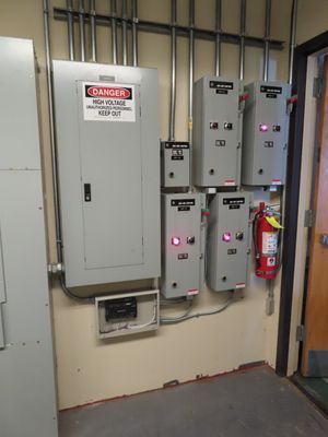 A wall-mounted array of electrical disconnects and breaker boxes in a commercial facility, including a red fire extinguisher for safety.
