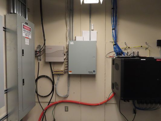A wall-mounted grey electrical sub-panel and communication box with a red cable connection in a mechanical room.