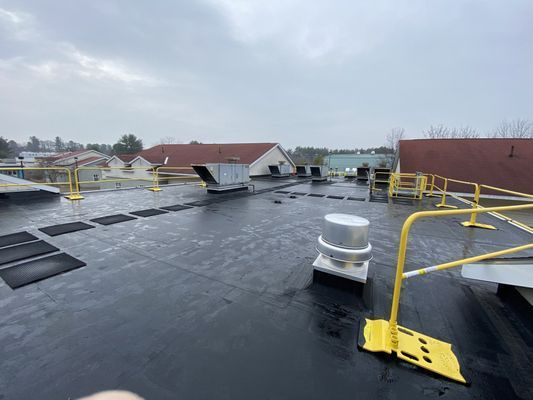 A flat black commercial rooftop featuring a large grey exhaust fan and yellow safety railings near the edge.