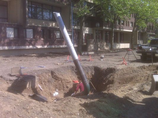 A large white-and-black industrial pipe being lowered into a deep excavation trench at a construction site.