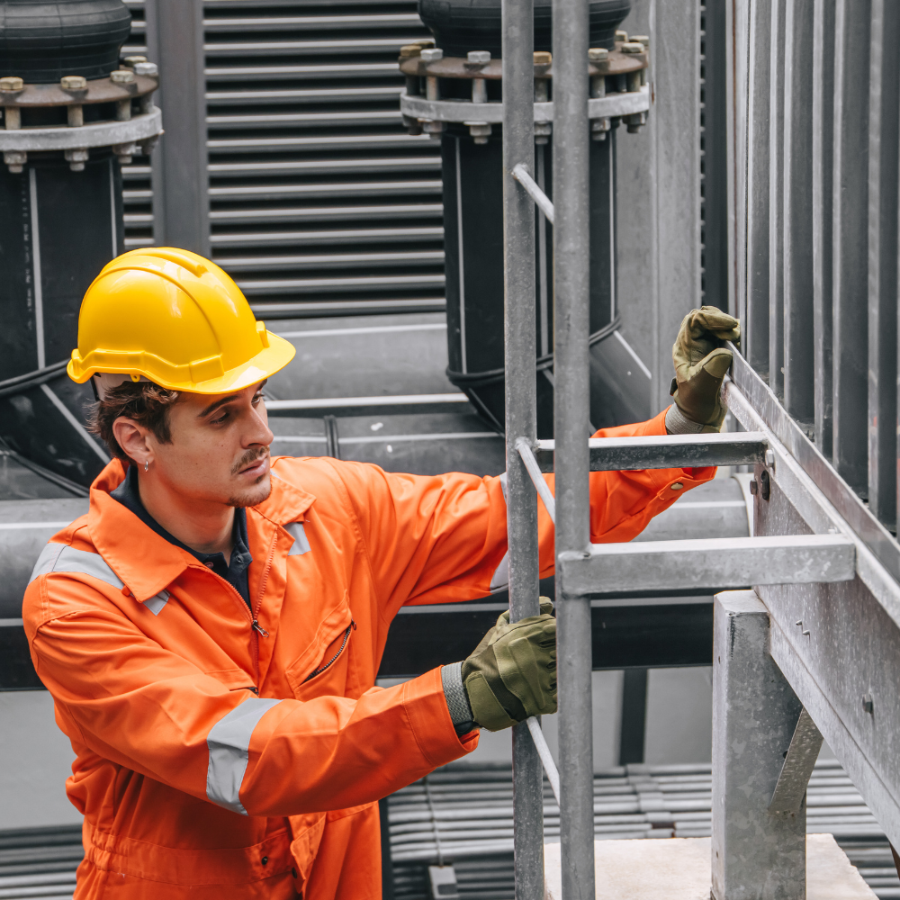A technician wearing an orange work suit and a yellow hard hat climbing a metal industrial ladder at a production facility.