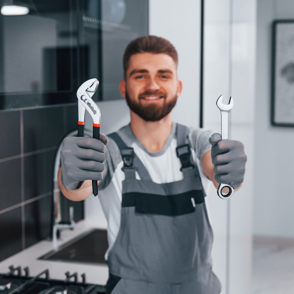 A smiling repairman wearing work gloves and grey overalls, holding up a pair of channel lock pliers and a combination wrench.