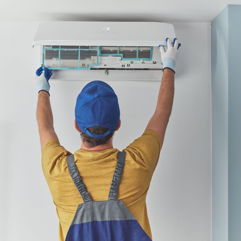 Technician in a blue cap removing or cleaning the air filter from a wall-mounted AC unit.