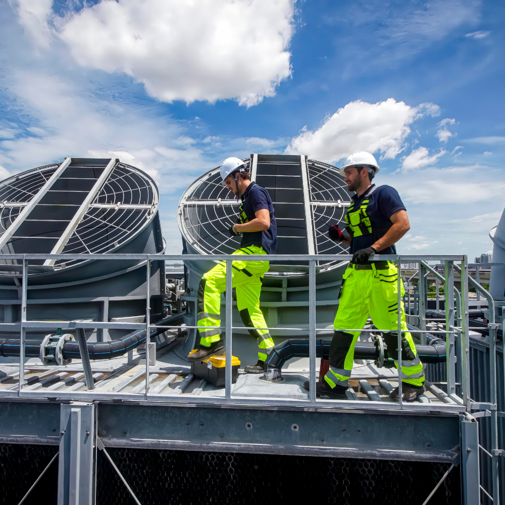 Two HVAC technicians in high-visibility safety uniforms and hard hats performing maintenance on large industrial cooling towers on a rooftop.