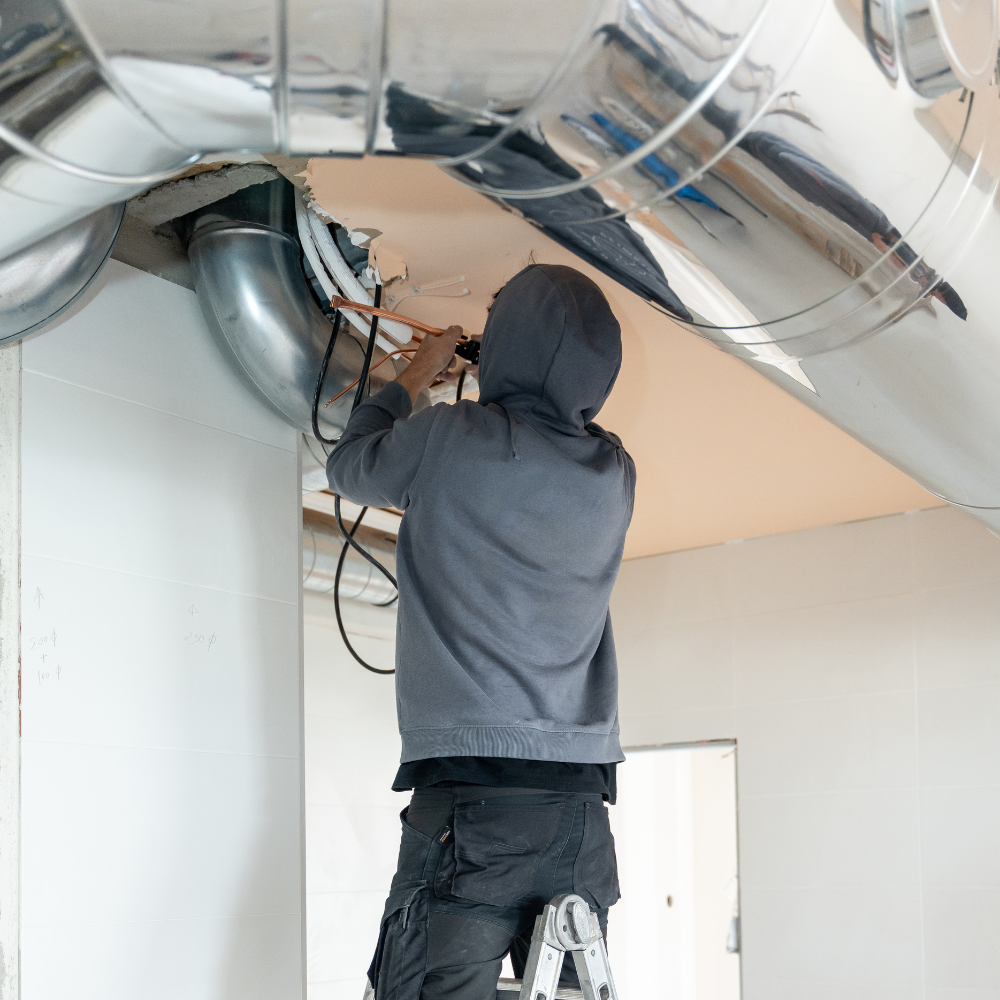 A worker in a grey hoodie reaching into a ceiling cavity to install or repair insulated ducting and electrical wiring.
