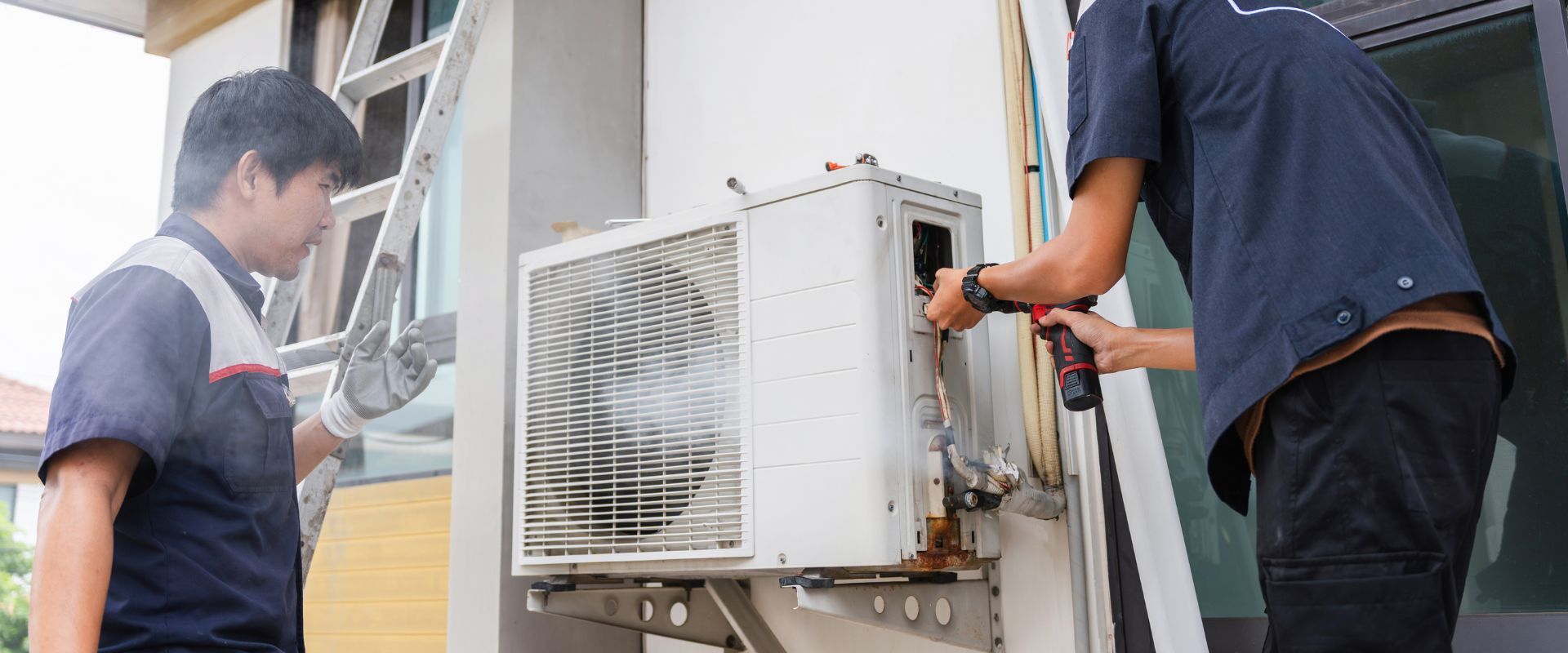 Two HVAC technicians collaborate to repair an outdoor condenser unit; one uses a power drill on the electrical panel while the other assists from a ladder.