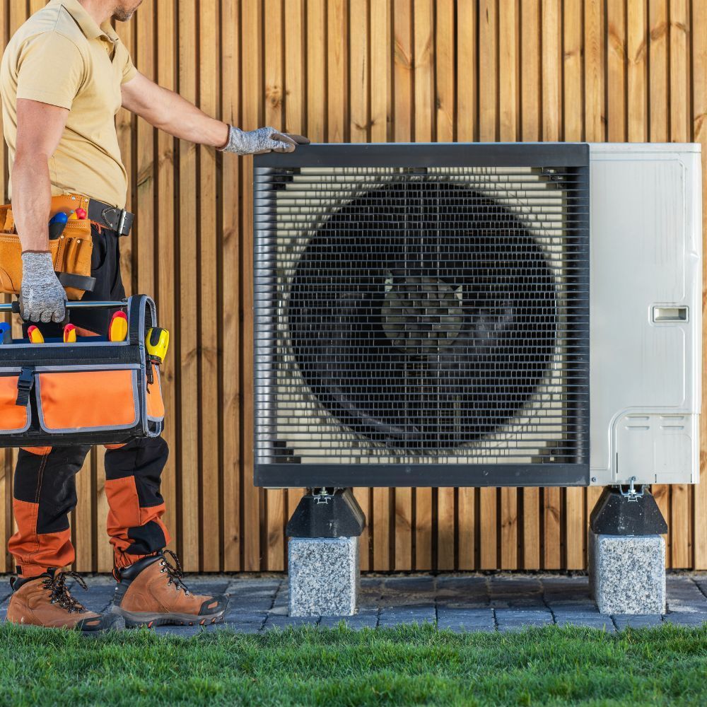 Technician carrying a tool bag while standing next to an outdoor heat pump unit mounted on concrete blocks.