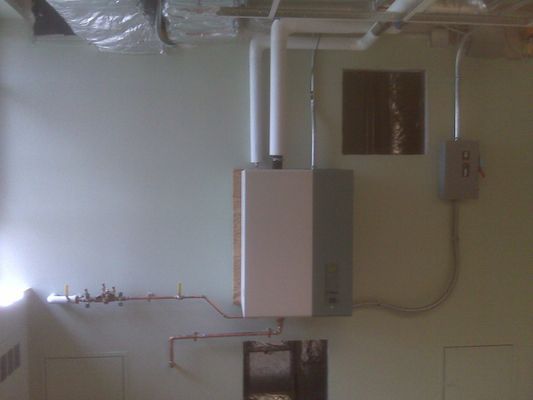 A grey wall-mounted industrial boiler or control box with copper piping and white insulated lines.