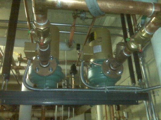 Close-up of industrial copper piping, brass valves, and green circulation pumps in a mechanical room.