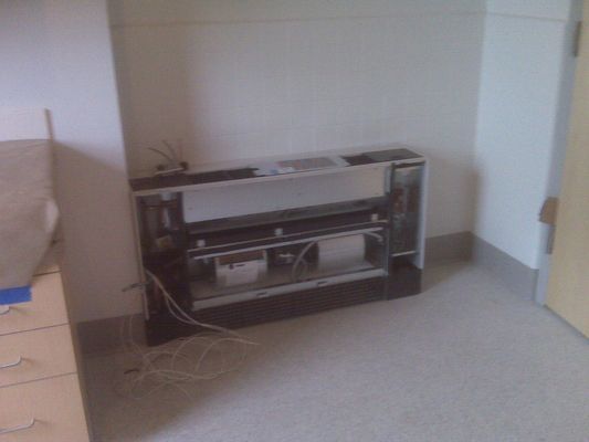 A partially disassembled white wall-mounted HVAC cabinet in a modern office or institutional room.