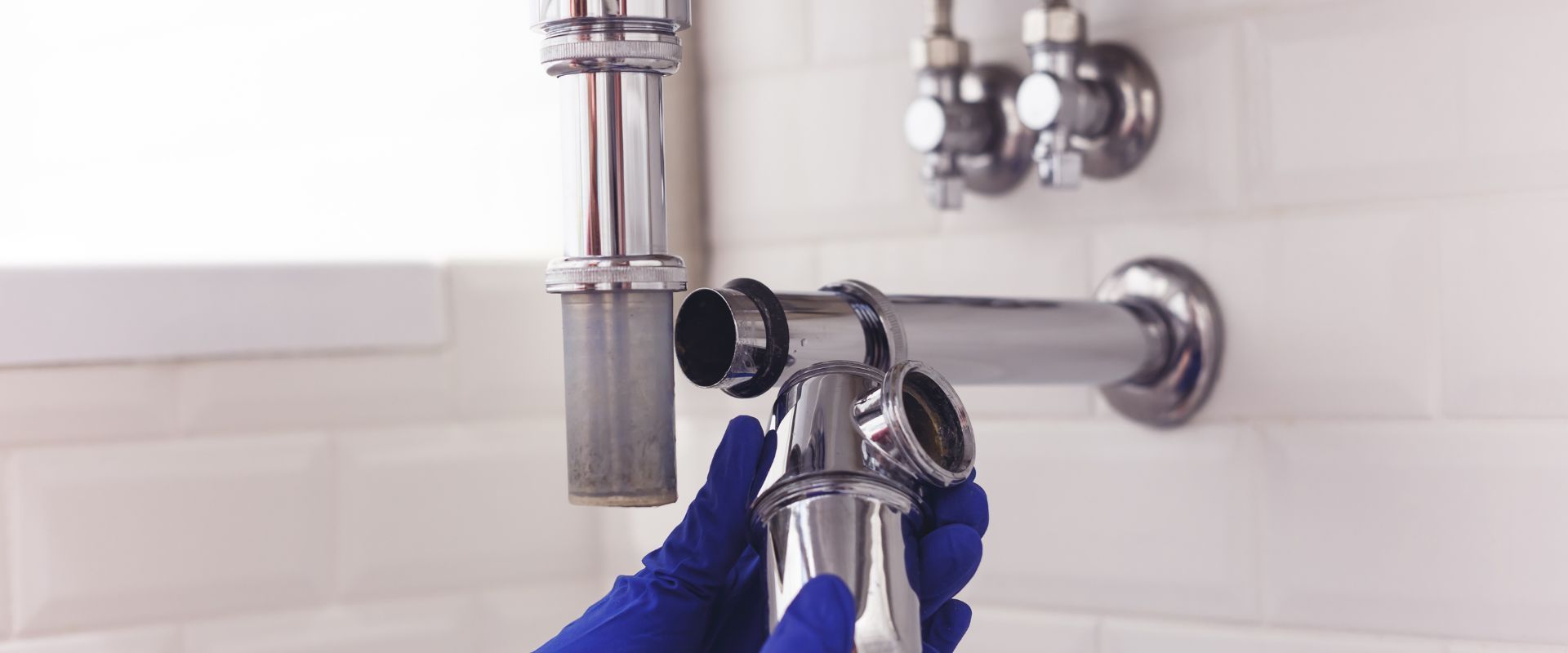 A close-up of a disassembled chrome drain pipe being inspected by a plumber wearing blue latex gloves.