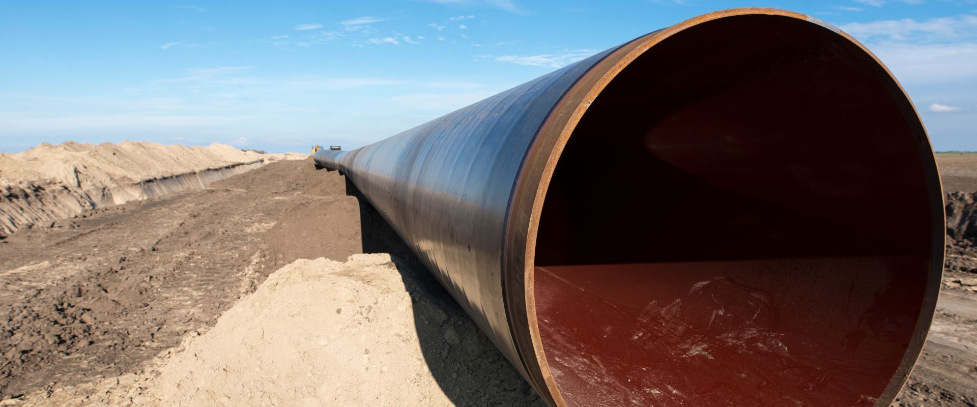 A perspective shot of a large-diameter steel pipe section showing the thick interior lining, extending across a flat construction site toward the horizon.