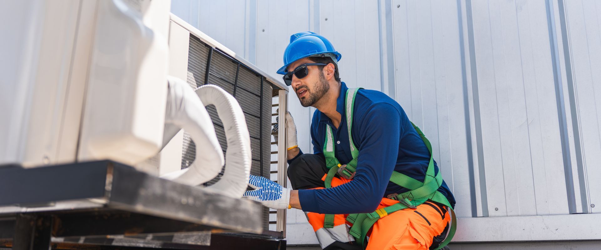 A technician wearing a blue hard hat, sunglasses, and a safety harness kneels down to perform a maintenance check on a large rooftop HVAC system.