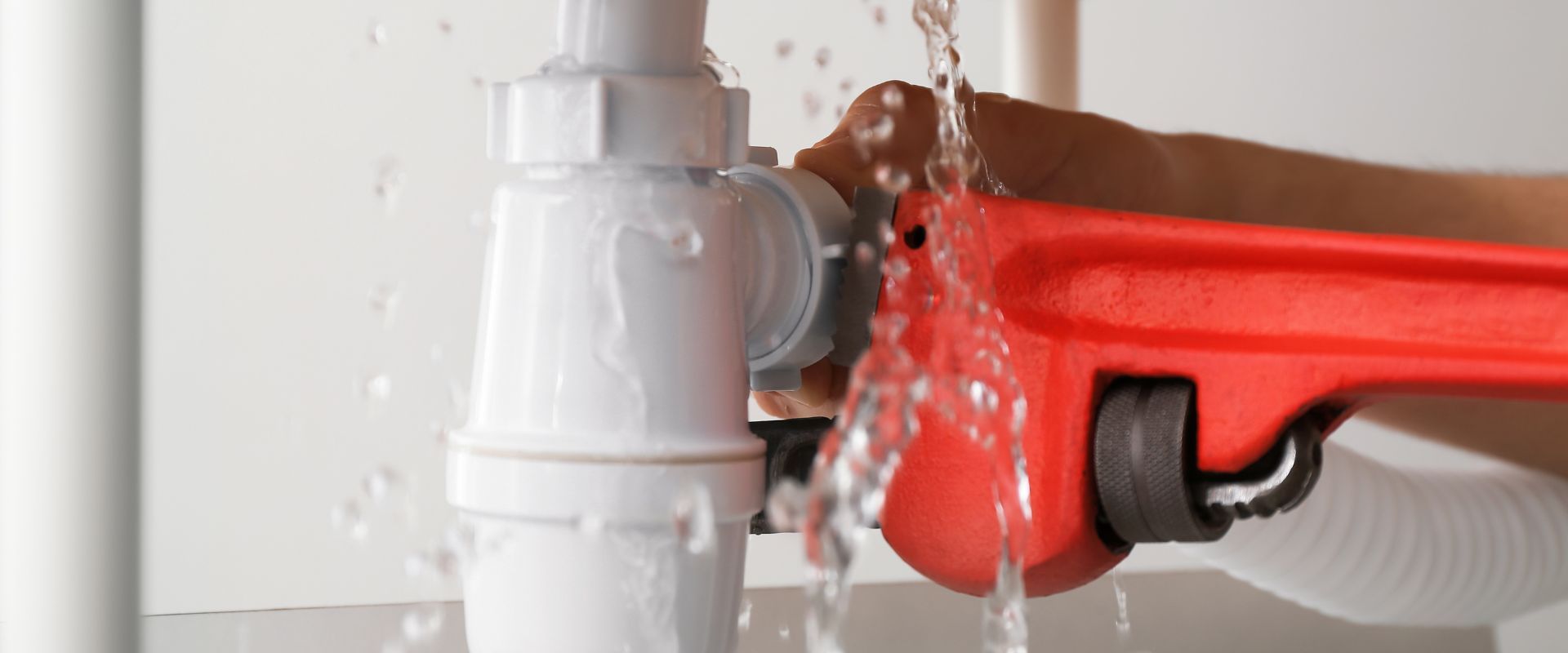 Water spraying from a leaking white plastic drain pipe as a technician attempts to secure the connection with a red pipe wrench.