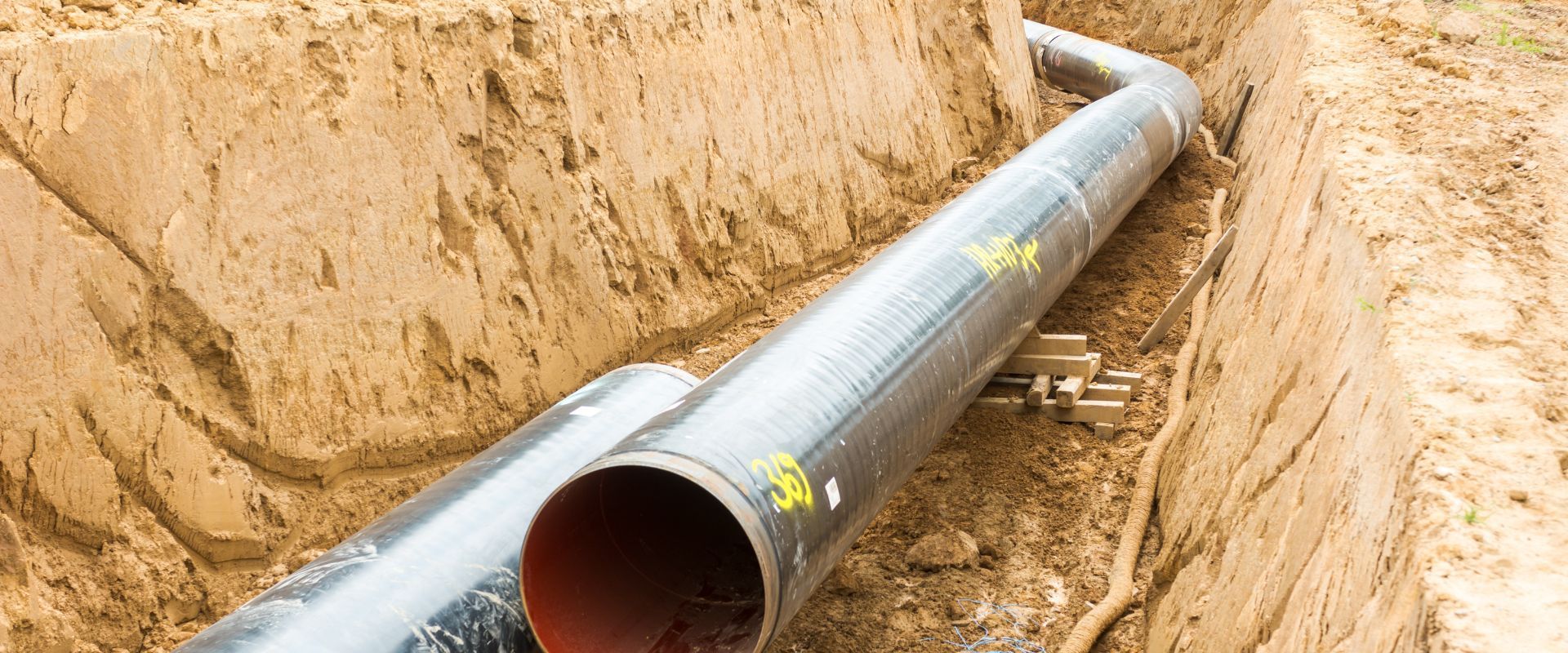 A large steel pipeline with a red-coated interior bending through a sandy trench, supported by wooden blocks during the installation phase.