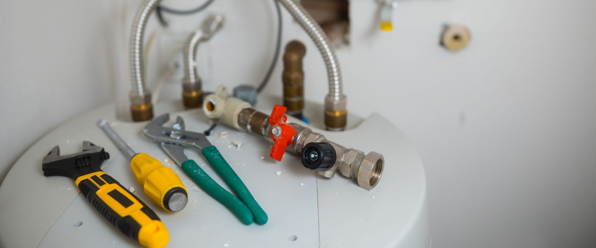 Various plumbing tools including pliers, a screwdriver, and a wrench lying next to a water heater unit and flexible metal hoses.