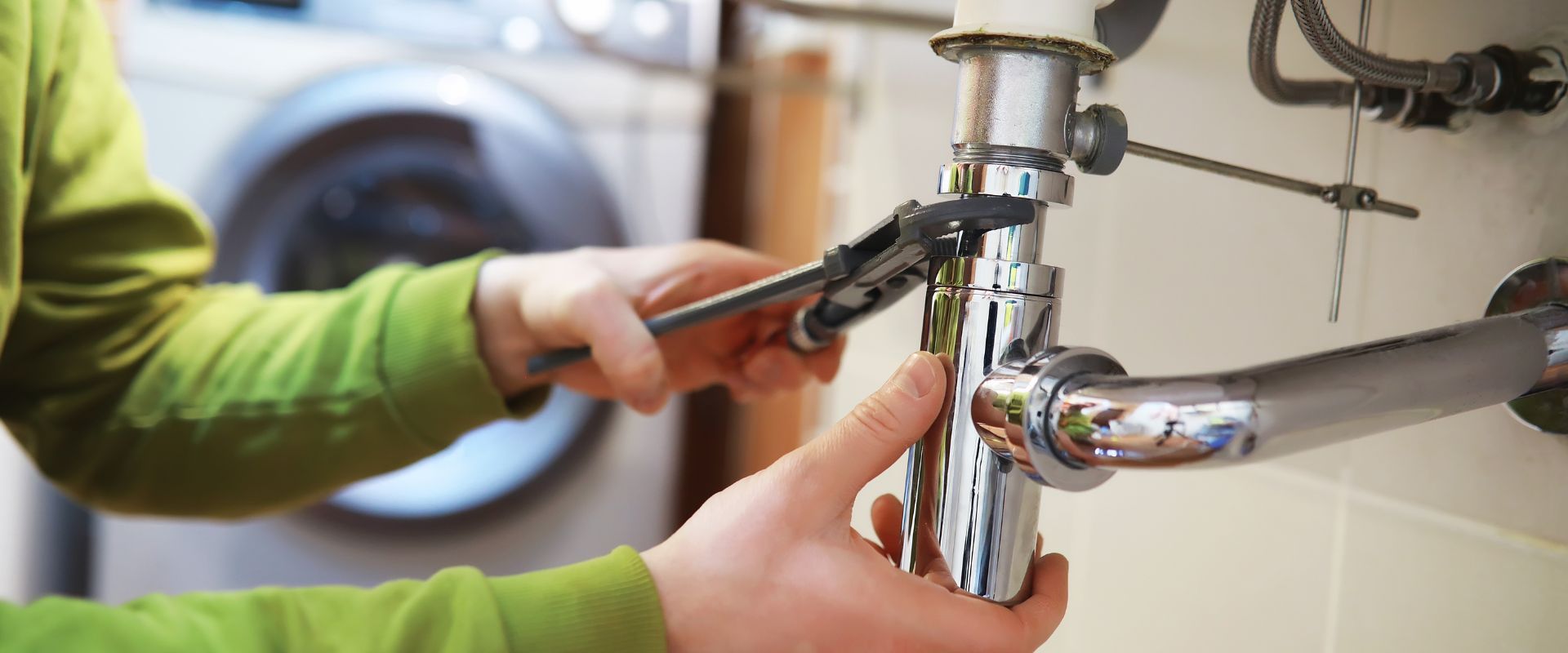 A plumber using a wrench to tighten a chrome p-trap assembly under a sink during a residential repair.