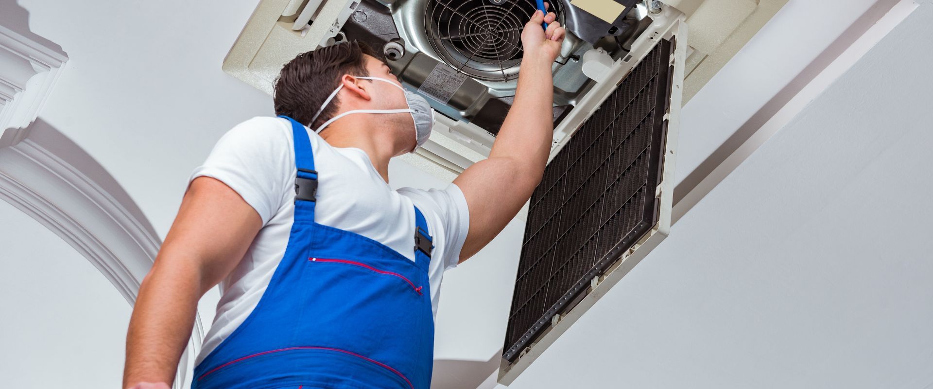 An HVAC professional in blue overalls and a face mask reaches up to inspect and clean the filter of a commercial ceiling-mounted air conditioning unit.