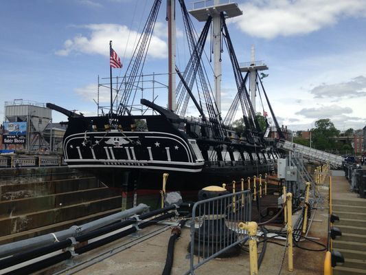 A historic wooden tall ship, the USS Constitution, docked in a dry dock for maintenance under a cloudy sky.