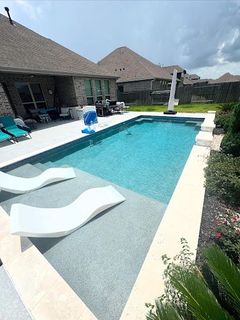Rectangular backyard pool with two white lounge chairs in the water. The pool has steps and is surrounded by a light-colored border.
