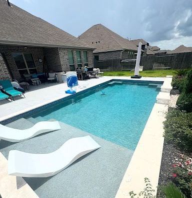 A rectangular swimming pool with clear blue water sits next to a patio with lounge chairs. A house is in the background.