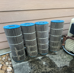 Four cylindrical pool filters with blue tops and a gray, pleated filter material, leaning against a wall on a concrete surface. They are covered in dirt.