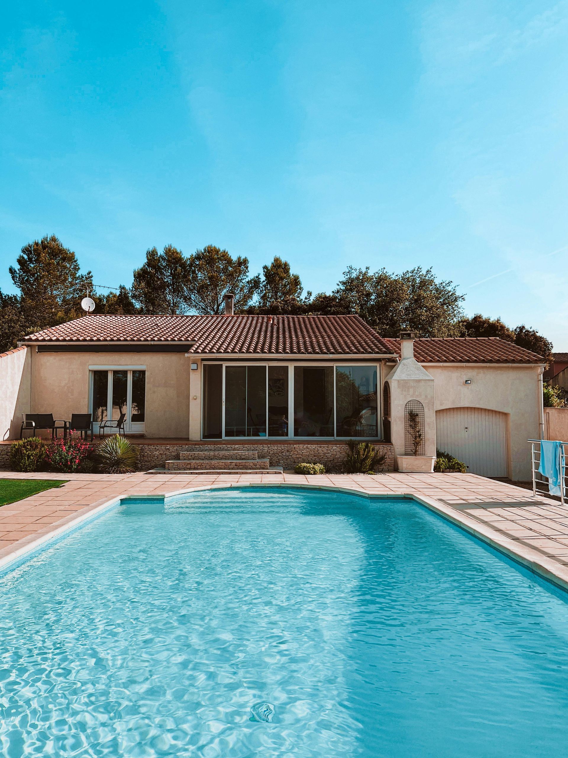 A swimming pool in front of a light-colored house with glass sliding doors. Blue sky overhead.