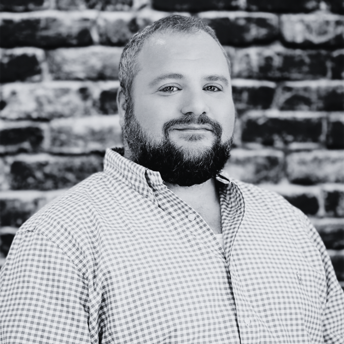 Man with a beard in a checkered shirt smiles in front of a brick wall.