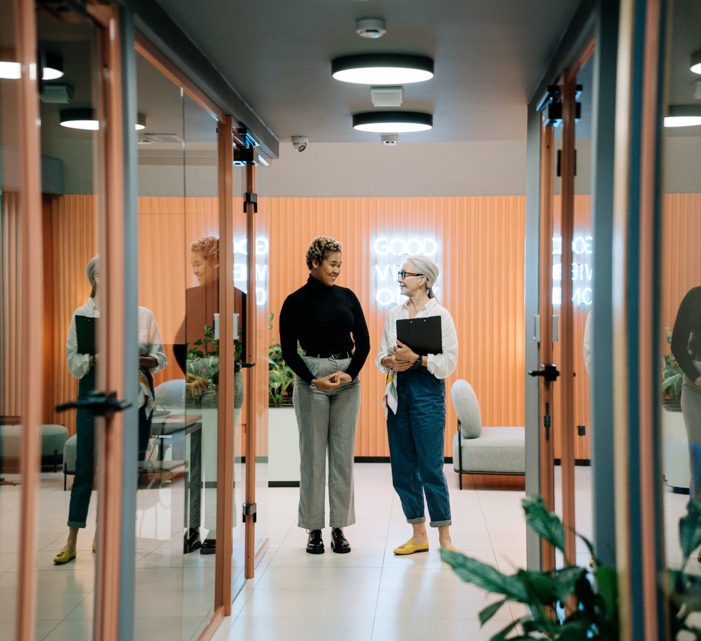 Two women standing in a hallway with a neon sign that says good work