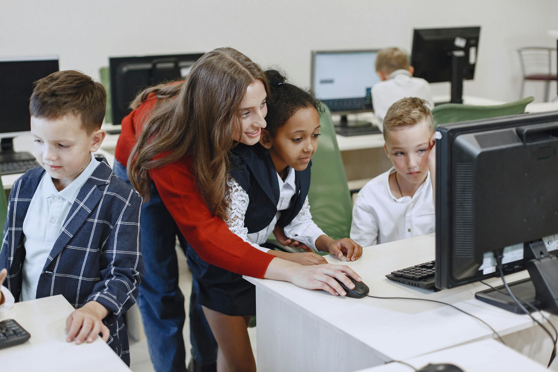 A teacher helps three students at a computer, one student on each side, in a classroom.