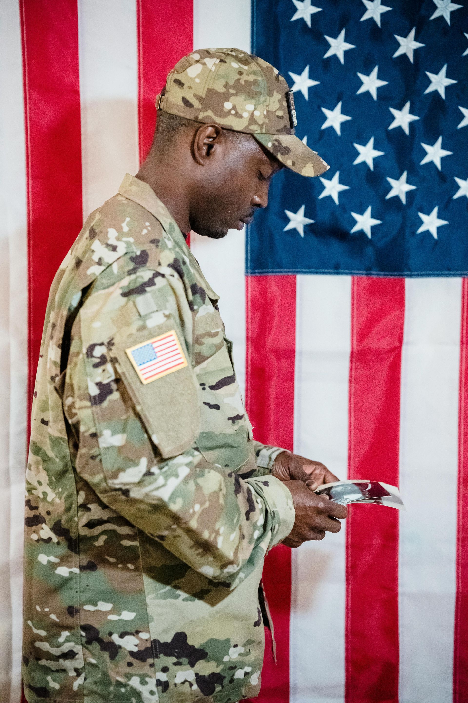 Soldier in camouflage uniform and cap, looking down at a small object, in front of an American flag.