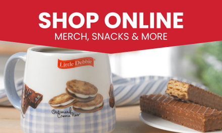 A mug with a picture of cookies on it is on a table next to a plate of food. Text is 