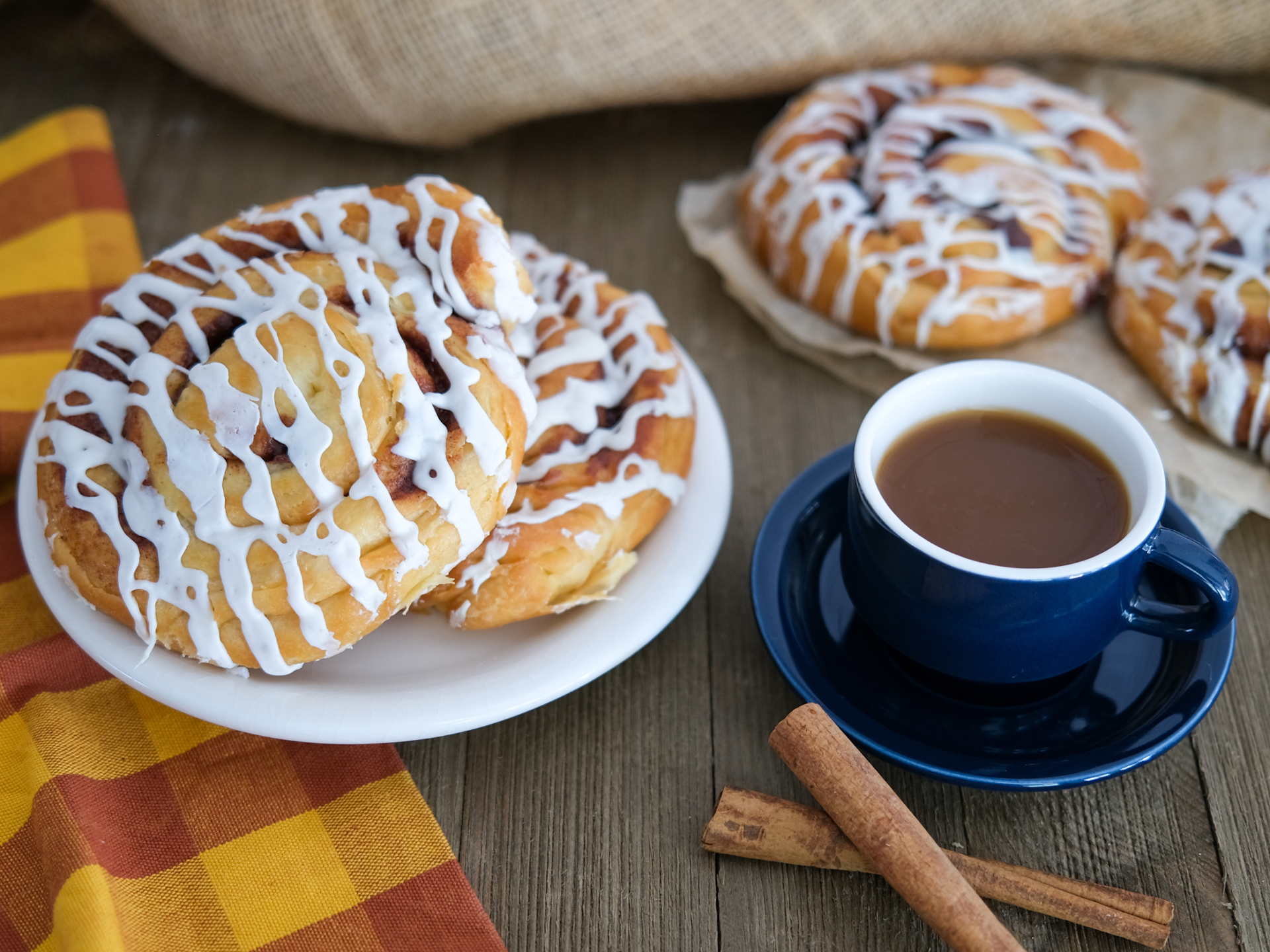 two cinnamon rolls on a white plate next two another cinnamon roll, a cup of coffee and cinnamon sticks