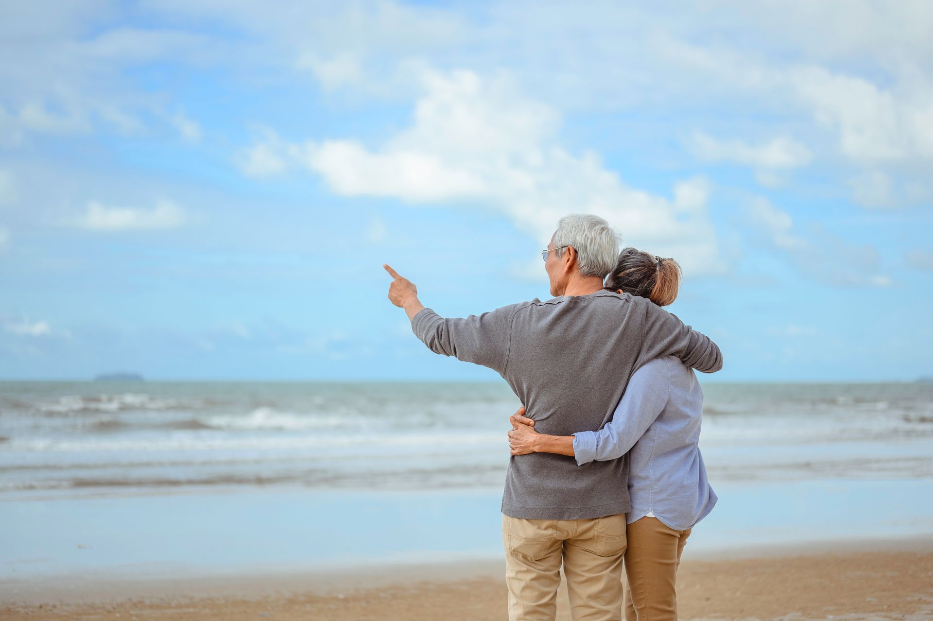 Couple with arms around each other at the beach; one points at the ocean under a blue sky.