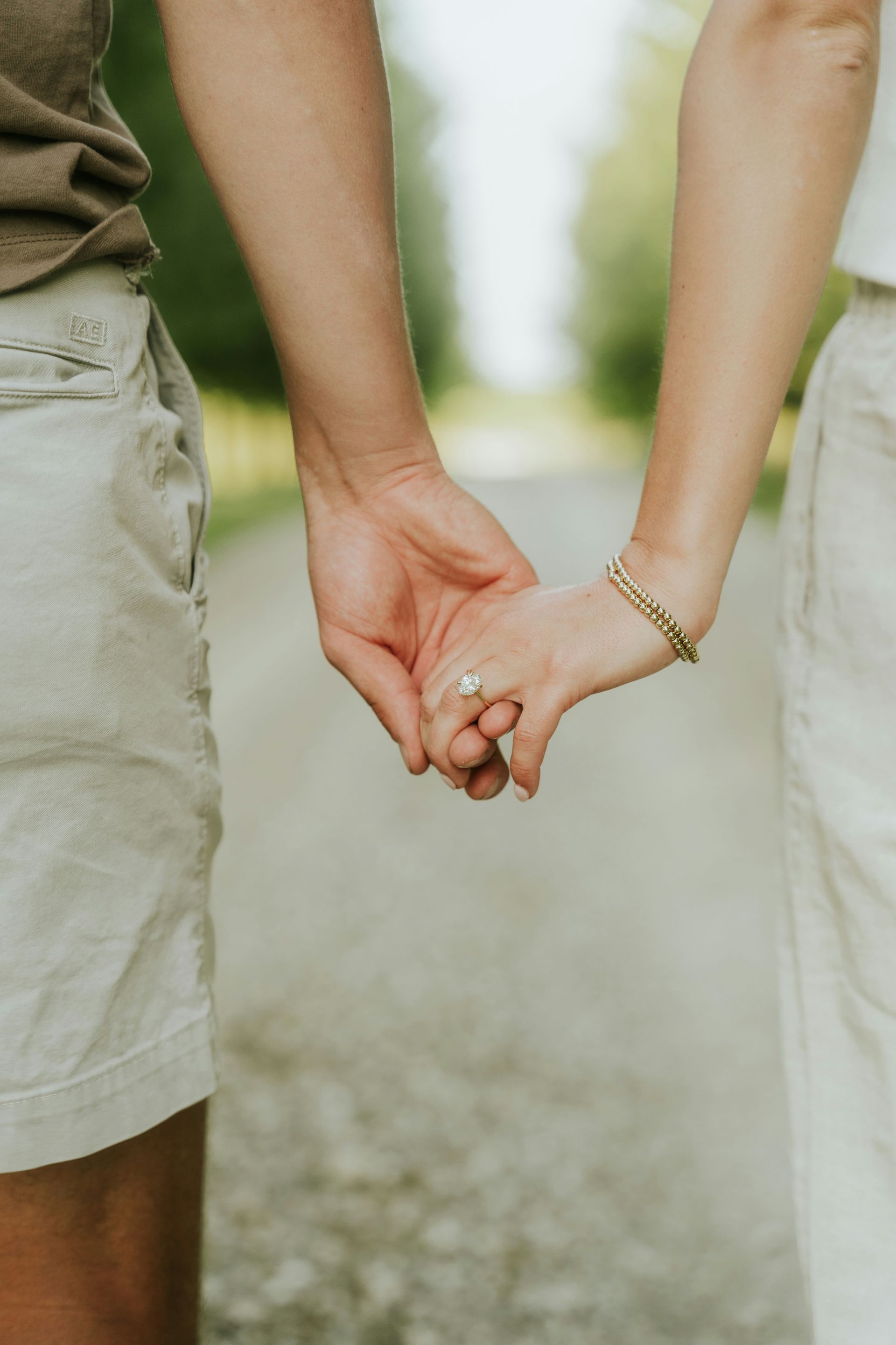 Couple holding hands on a path, wearing shorts and dress. Engagement ring visible.