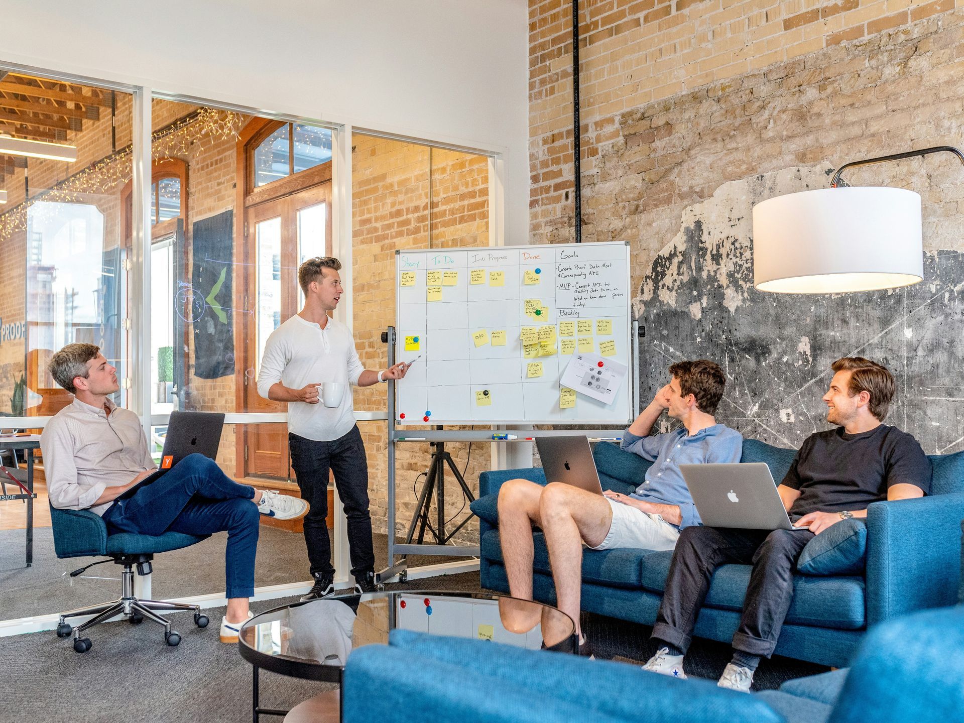 Four men in a modern office during a presentation, one standing at a whiteboard, the others using laptops and listening.