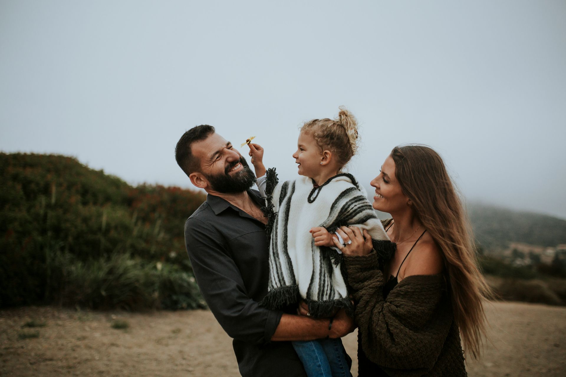 Family on a beach laughing; father holding child, mother smiling; overcast day.