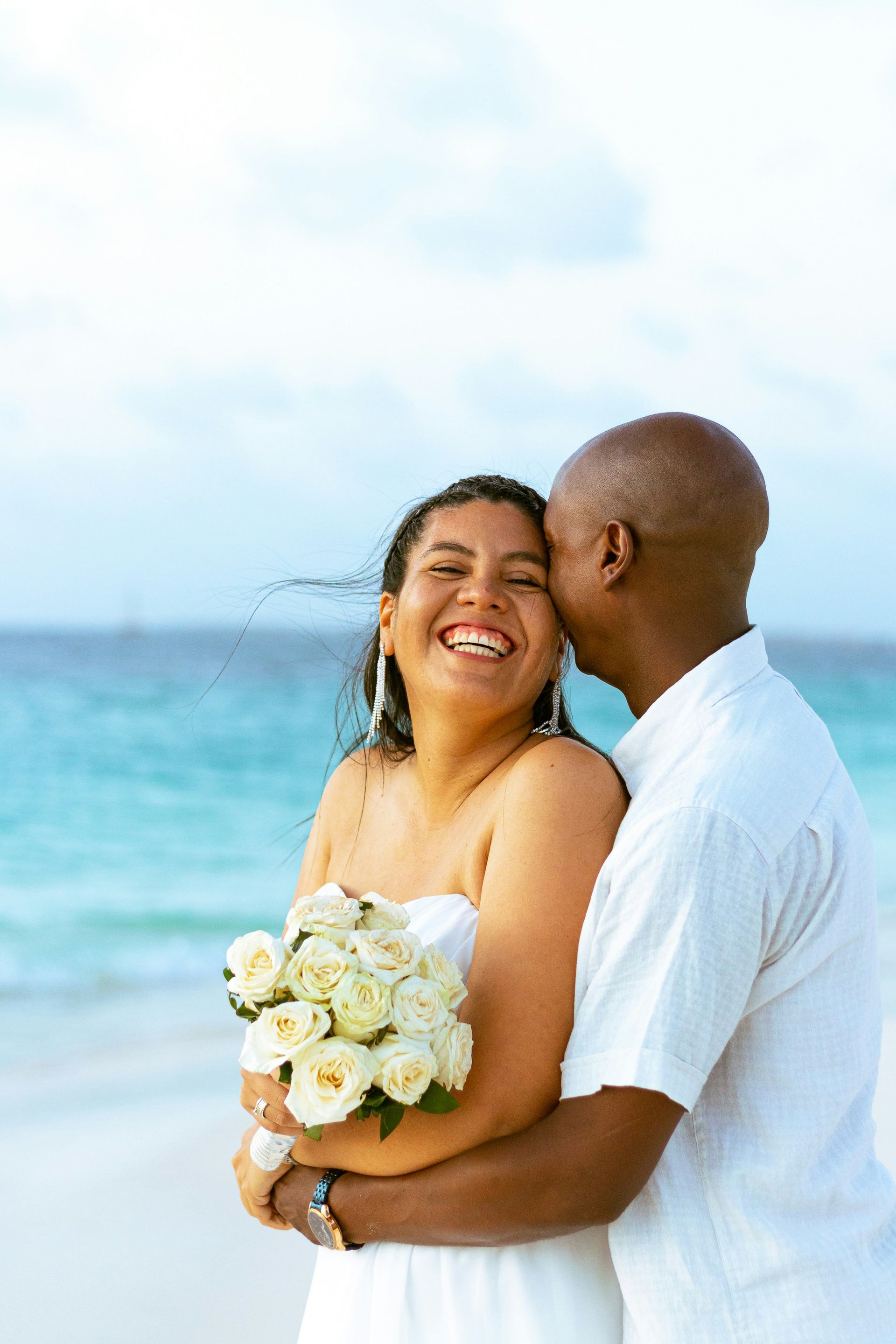Couple on a beach embracing; woman smiles holding bouquet, man kisses her cheek; ocean backdrop.