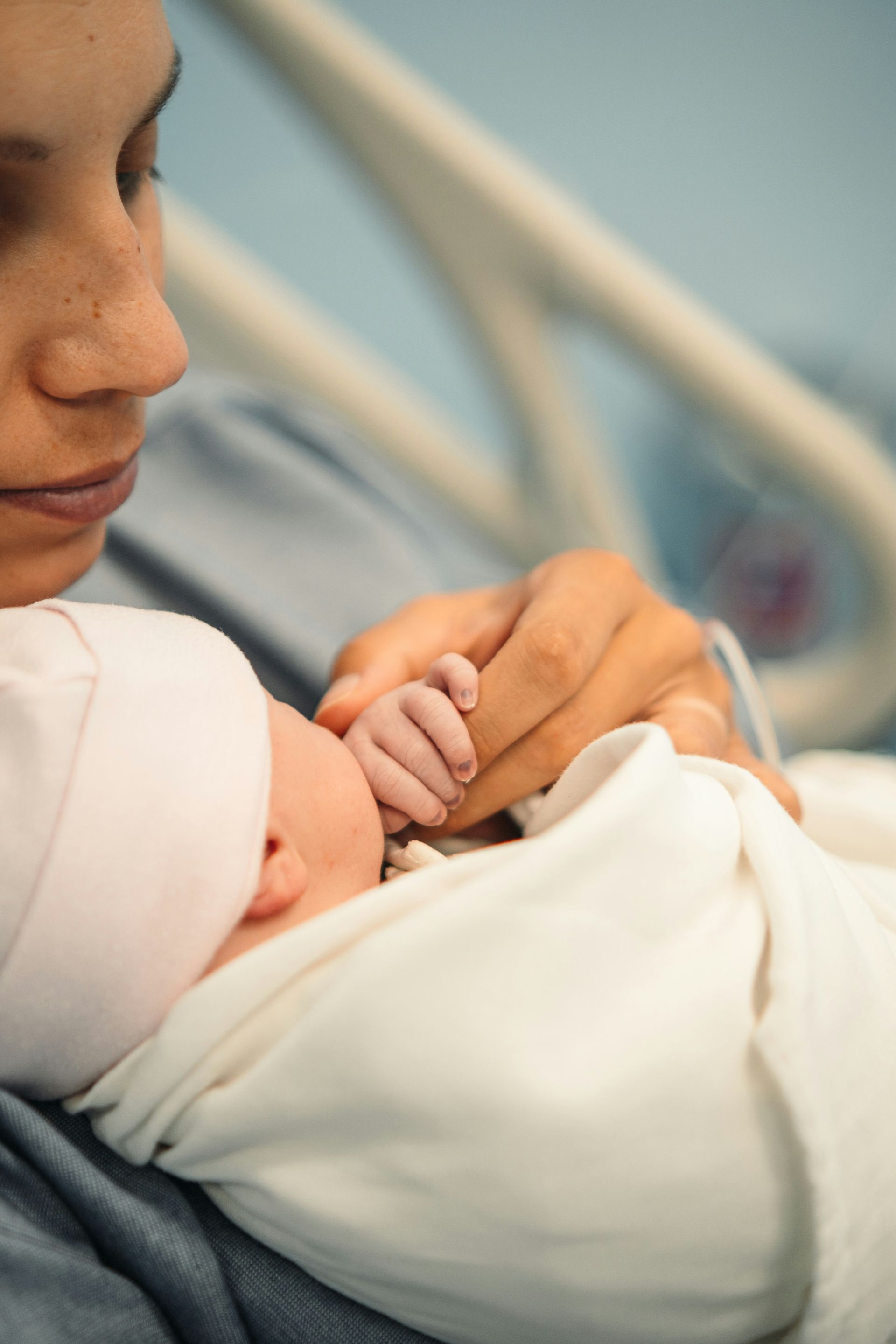 Mother holds newborn baby in a hospital. The baby's hand gently clasps mother's finger.