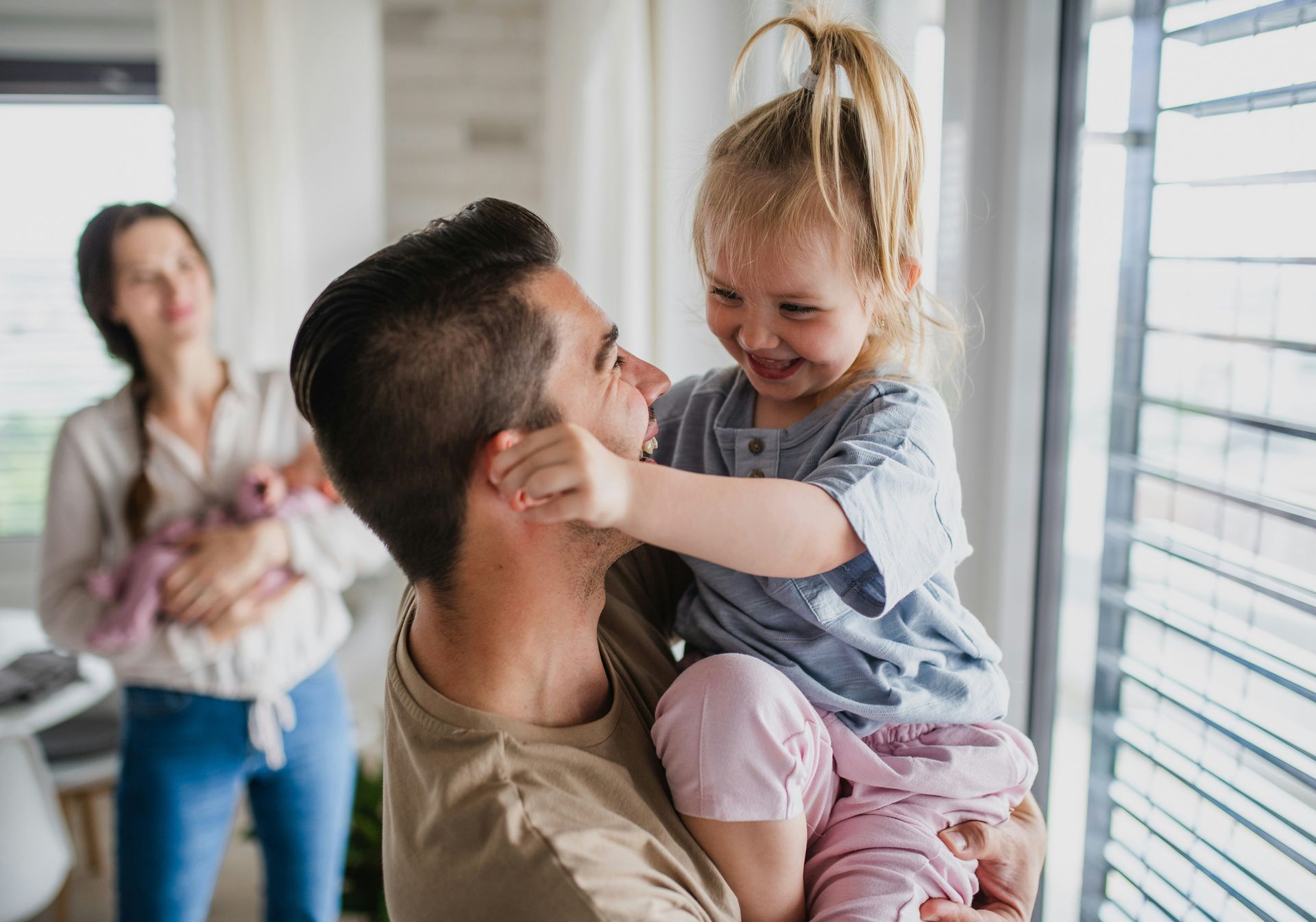 Father holding and smiling at a child, with mother holding an infant in the background.