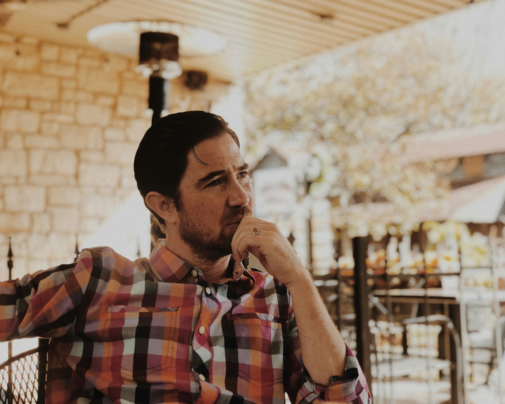 Man in plaid shirt, thoughtful expression, outdoors with stone wall, hand to his chin.