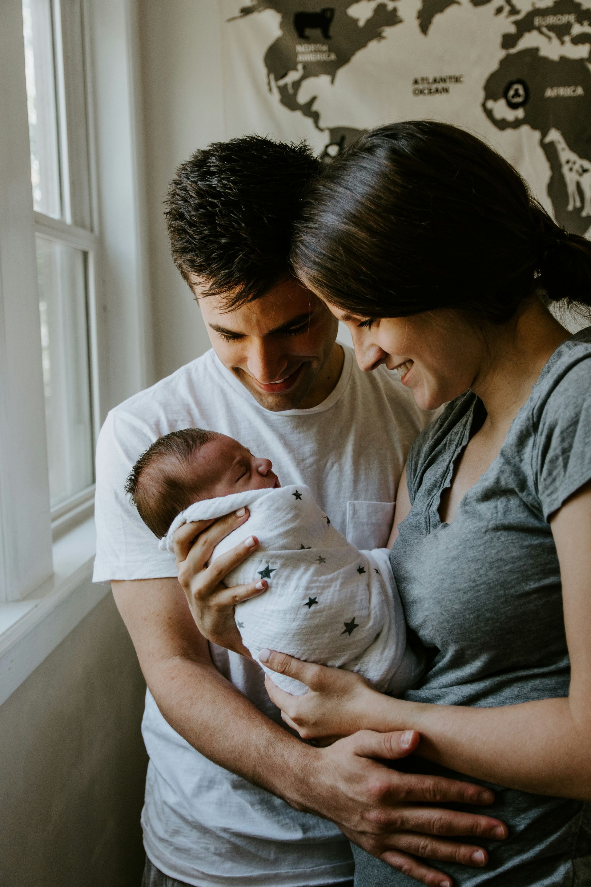 Couple gazing at a newborn baby wrapped in a swaddle blanket near a window.