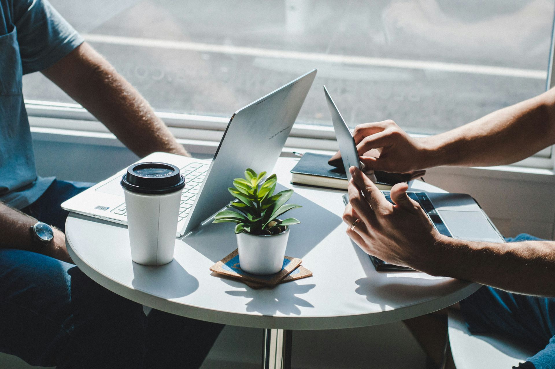 Two people working on laptops at a small table, next to a window; coffee cup and plant are also on the table.