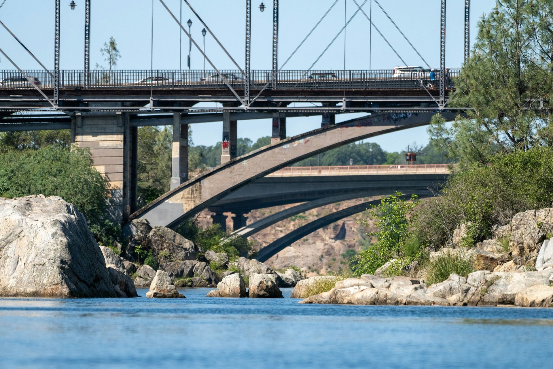 Three bridges over a river with rocks in foreground; blue water, and green foliage.